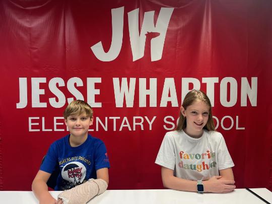 Two Jesse Wharton Elementary Students sit in front of a red school banner