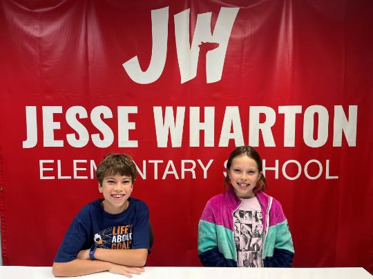 Two Jesse Wharton Elementary students sit in front of a red backdrop with their school name on it